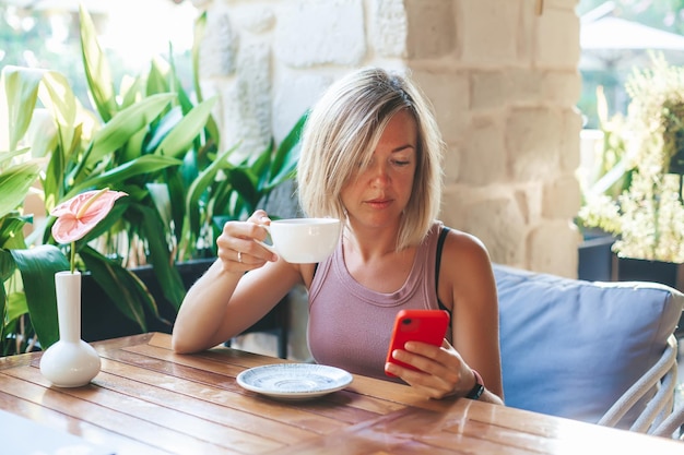 Mujer relajada sosteniendo y usando un teléfono inteligente para mensajes sms, escribiendo un teléfono celular con pantalla táctil en el café soleado, bebiendo té. Estilo de vida, tecnología y concepto de red de medios sociales.