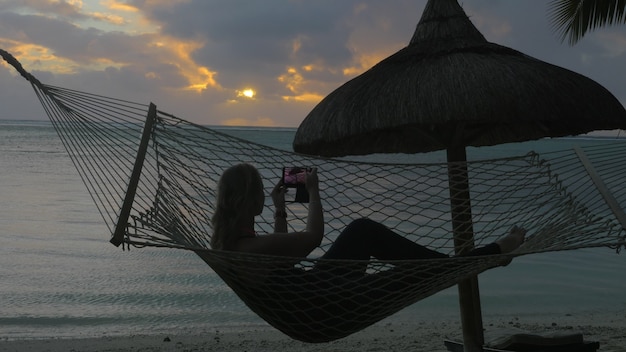 Mujer relajándose en una hamaca en la playa y tomando fotos de la celda