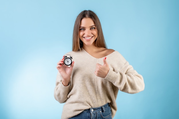 Mujer con reloj despertador mostrando pulgares arriba aislado sobre azul