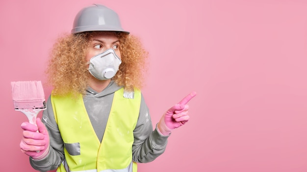 Mujer reparadora o pintora indica distancia en el espacio de copia tiene pincel de pintura ocupado haciendo renovación de la casa lleva casco y uniforme de protección respiratoria