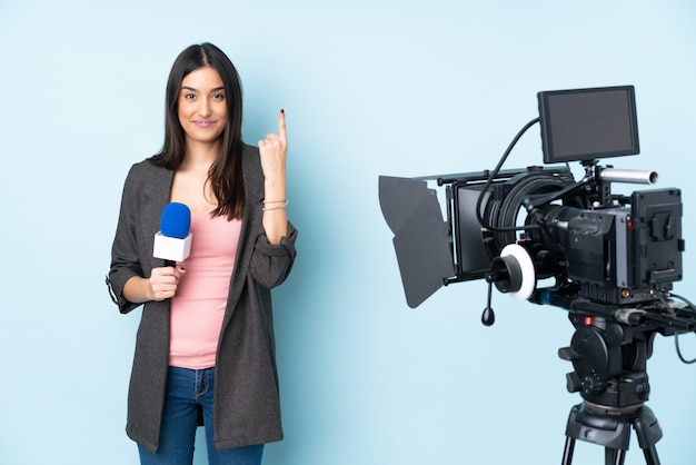 Mujer reportera sosteniendo un micrófono y reportando noticias en la pared azul señalando con el dedo índice una gran idea