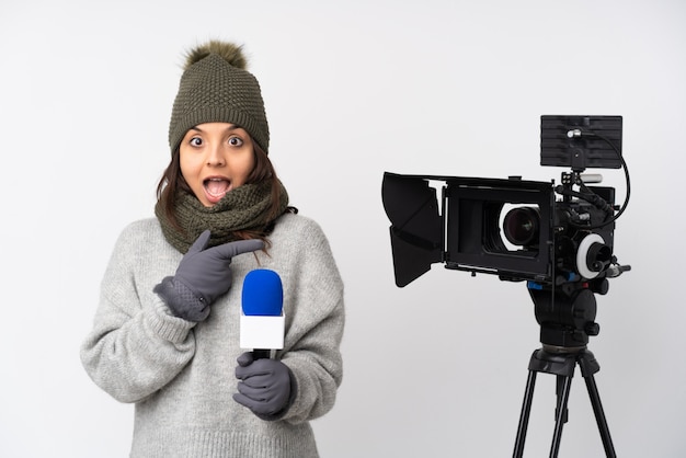 Mujer reportera sosteniendo un micrófono y reportando noticias sobre una pared blanca sorprendida y apuntando con el dedo a un lado