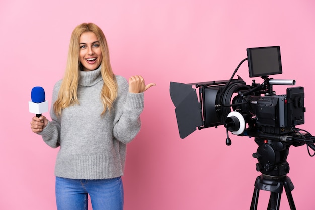 Mujer reportera sosteniendo un micrófono y reportando noticias sobre una pared rosa aislada apuntando hacia un lado para presentar un producto