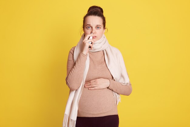 Mujer con resfriado durante el embarazo que usa spray nasal para aliviar los síntomas, futura mamá enferma, con mirada triste y poco saludable, posa aislada sobre una pared amarilla.