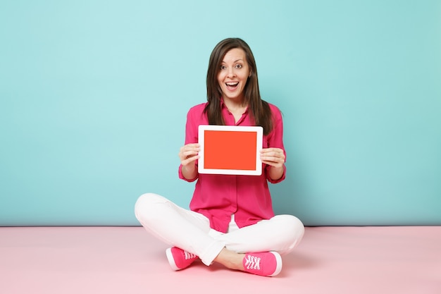 Mujer de retrato de cuerpo entero en blusa de camisa rosa, pantalón blanco sentado en el piso con tablet pc aislado en la pared pastel azul rosa brillante.