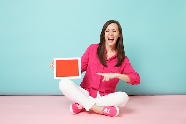 Mujer de retrato de cuerpo entero en blusa de camisa rosa, pantalón blanco sentado en el piso con tablet pc aislado en la pared pastel azul rosa brillante.