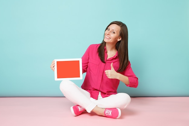 Mujer de retrato de cuerpo entero en blusa de camisa rosa, pantalón blanco sentado en el piso con tablet pc aislado en la pared pastel azul rosa brillante.