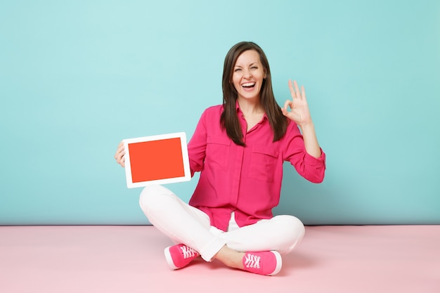 Mujer de retrato de cuerpo entero en blusa de camisa rosa, pantalón blanco sentado en el piso con tablet pc aislado en la pared pastel azul rosa brillante.