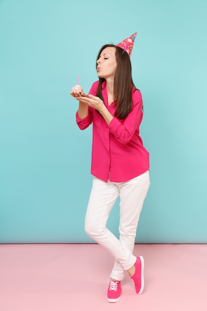 Mujer de retrato de cuerpo entero en blusa de camisa rosa, pantalón blanco, sombrero de cumpleaños con cupcake posando aislado en la pared pastel azul rosa brillante.