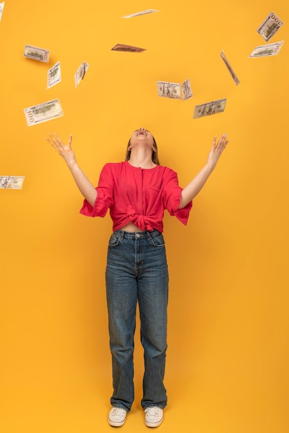 Mujer riendo feliz lanzando billetes de dólar mientras está de pie contra el fondo naranja Abundancia y desperdicio de dinero Marco vertical