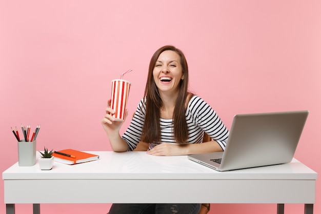 Mujer riendo joven que sostiene la taza plástica con cola o refresco sentarse y trabajar en el escritorio blanco con computadora portátil contemporánea aislada sobre fondo rosa pastel. Concepto de carrera empresarial de logro. Copie el espacio.