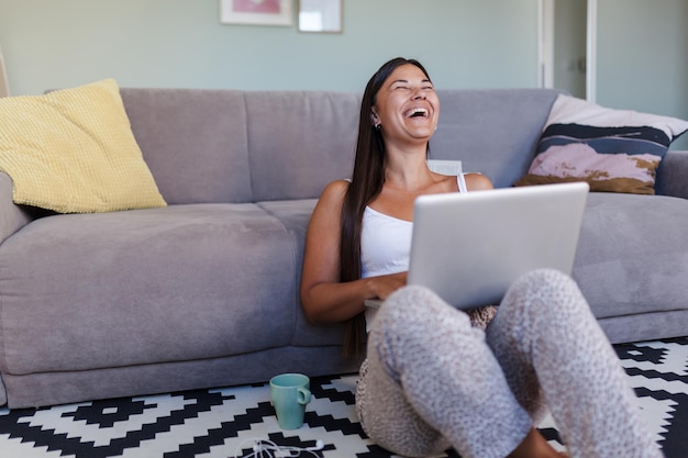 Mujer riendo usando una computadora portátil mientras está sentada en el sofá en casa