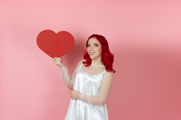 mujer riendo con un vestido blanco y con el pelo rojo abraza un gran corazón rojo de papel