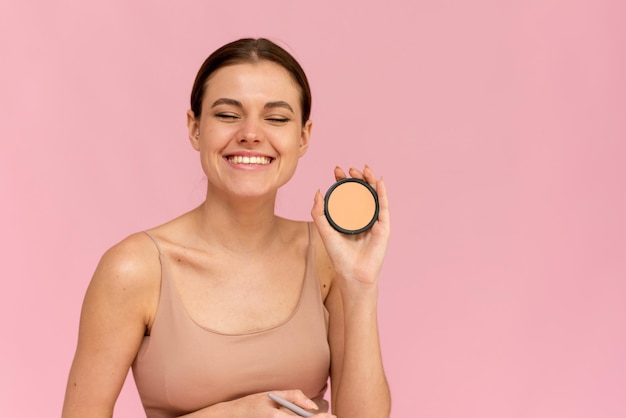 Foto mujer riéndose aplicando polvo en la cara sobre un fondo rosa en el estudio.