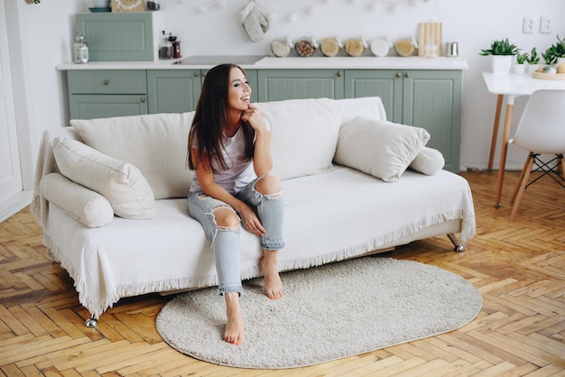 Mujer en Ripped Jeans se sienta en un estado de ánimo alegre en el sofá Inicio interior acogedor Cocina verde en segundo plano.