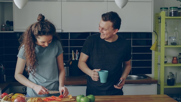 Mujer rizada cerca de la mesa cortando pimiento rojo en la cocina en casa, su novio le muestra algo divertido en el teléfono inteligente y están sonriendo