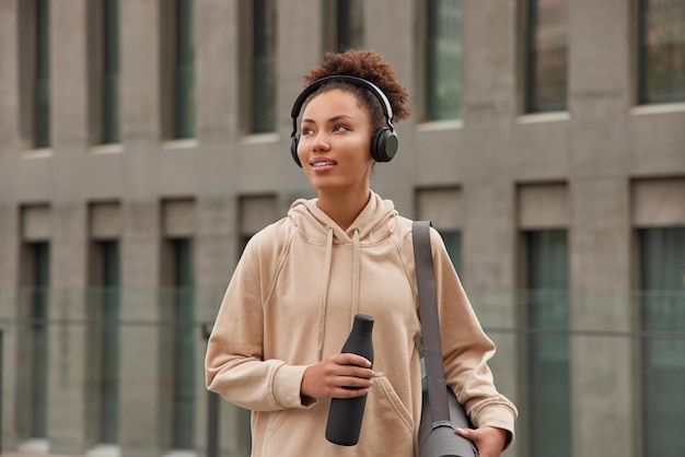 Mujer rizada deportiva pensativa vestida con capucha beige pasea al aire libre contra un edificio moderno escucha música a través de auriculares sostiene una botella de agua lleva karemat enrollado lleva un estilo de vida saludable