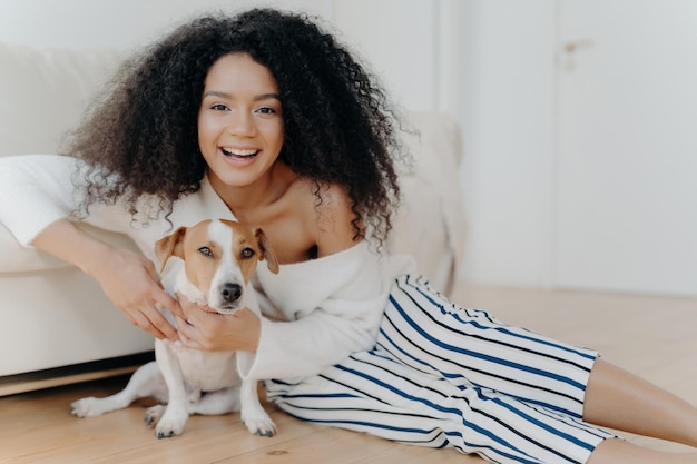 Mujer rizada feliz con ropa de moda juega con un perrito divertido en el suelo compartiendo momentos alegres y amor