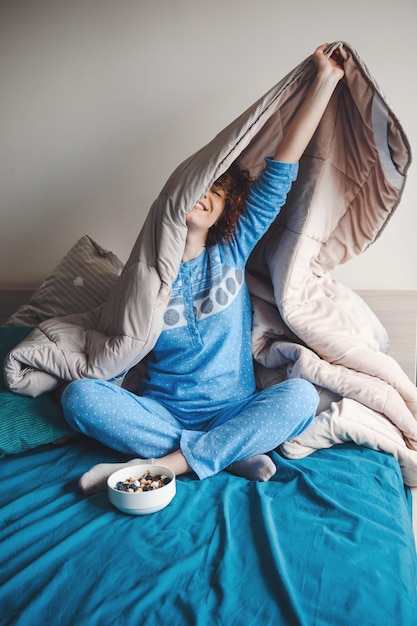 Mujer rizada que pasa su mañana de fin de semana en la cama debajo de una manta vistiendo un pijama azul saludable me ...