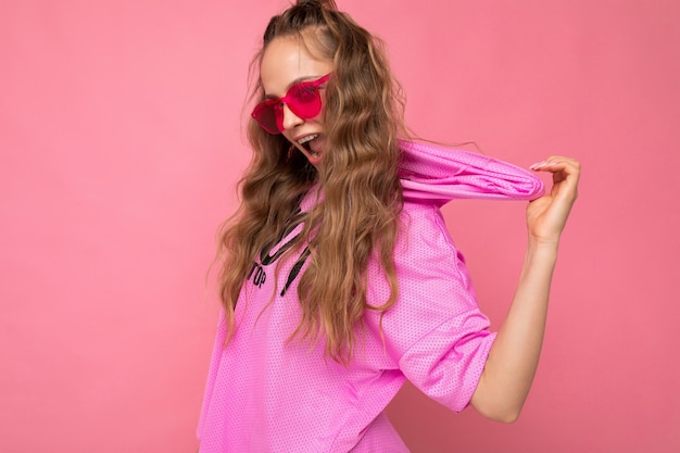 Mujer rizada rubia joven divertida bastante positiva aislada sobre la pared del fondo rosado que lleva la camiseta rosada ocasional y las gafas de sol coloridas elegantes que miran la cámara y que se divierten