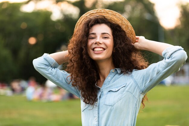 Foto mujer rizada con sombrero de paja riendo, mientras camina en el parque verde