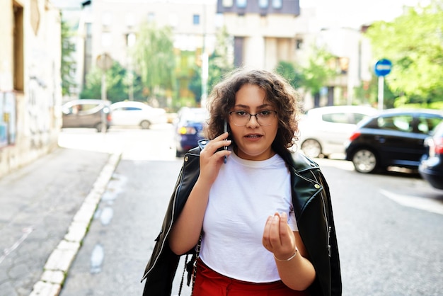 Mujer rizada sonriente con gafas de sol de moda camina por la calle central de la ciudad y usa su teléfono