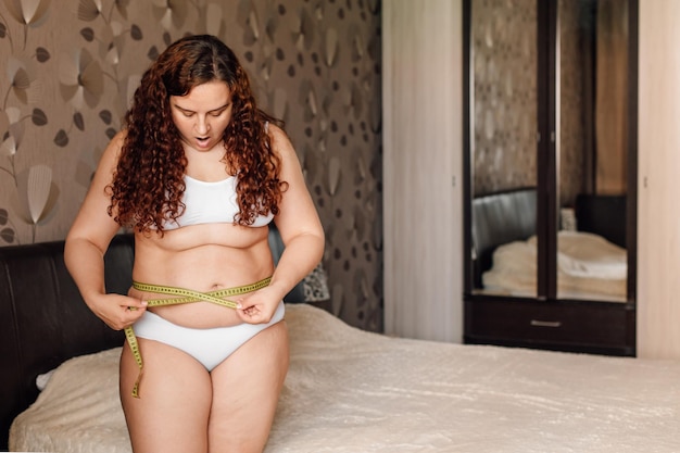 Mujer rizada de talla grande en ropa interior blanca parada cerca de la cama en el dormitorio mide la circunferencia de la cintura con cinta adhesiva y sorprende Las niñas con sobrepeso luchan contra la obesidad Control corporal del programa de pérdida de peso