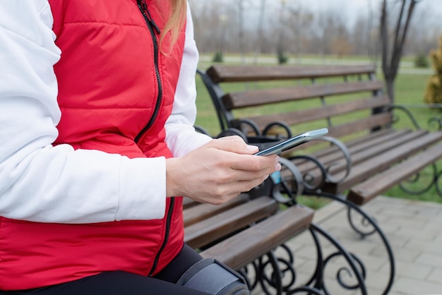 Mujer con rodillera u ortesis después de una cirugía de pierna sentada en el parque usando un teléfono inteligente