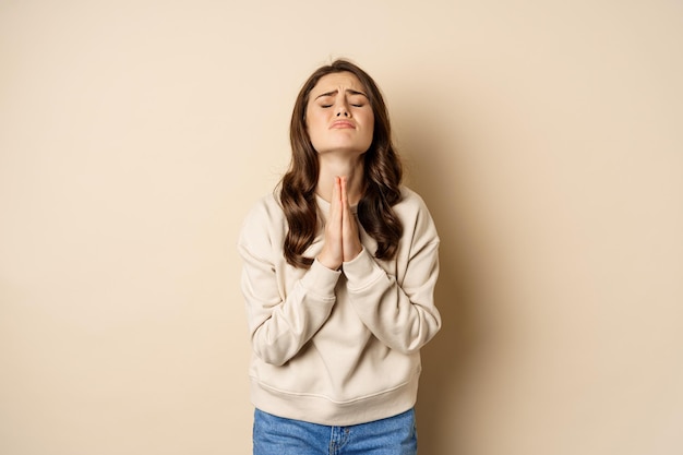 Mujer rogando y suplicando a Dios suplicando decir por favor de pie desesperada sobre fondo beige