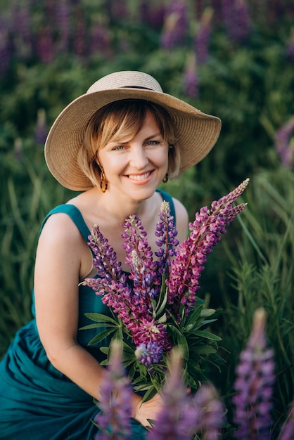 Mujer romántica feliz que sonríe en un vestido azul y un sombrero en un campo con un ramo de flores púrpuras de altramuces.