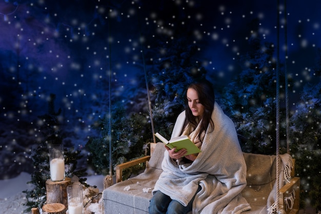 Mujer romántica leyendo un libro mientras está sentada en un columpio y envuelta en una manta cálida por la noche en un parque cubierto de nieve con abetos, mientras nieva