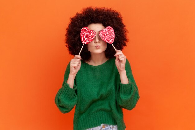 Foto mujer romántica con peinado afro con suéter verde de estilo casual que cubre sus ojos con dulces en forma de corazón y labios fruncidos haciendo gesto de beso estudio interior aislado en fondo naranja