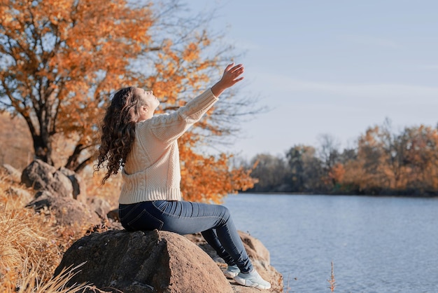 Mujer romántica pensativa sentada en las rocas en la orilla del río al atardecer en el día de otoño