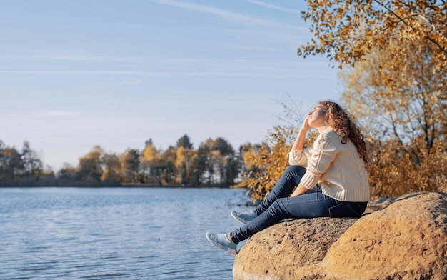 Mujer romántica pensativa sentada en las rocas en la orilla del río al atardecer en el día de otoño