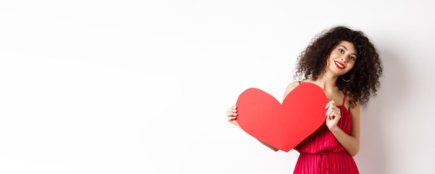 Mujer romántica vestida mostrando un gran corazón rojo enamorándose sonriendo feliz a la cámara con fondo blanco