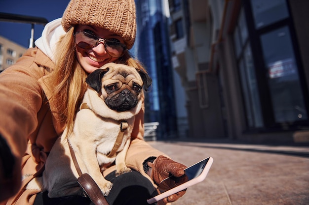 Foto una mujer con ropa abrigada se hace selfie con su pequeño perro pug cerca del edificio comercial que está en el fondo.