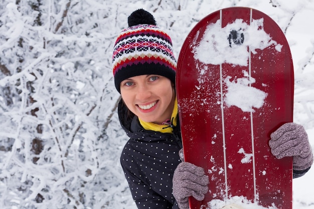 Mujer en ropa de abrigo sonríe y sostiene una tabla de snowboard en el fondo de un paisaje invernal