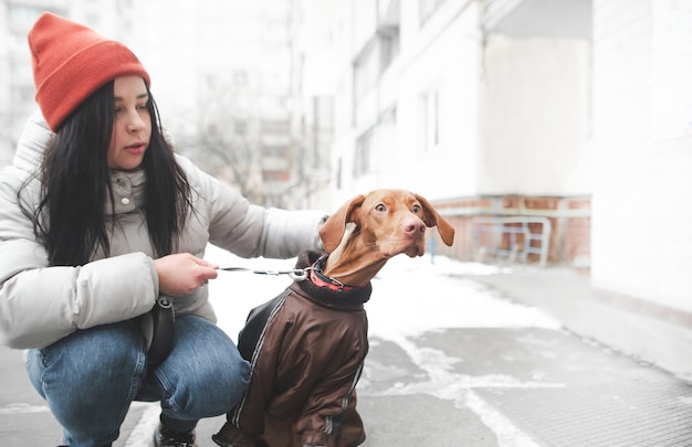 Mujer en ropa de abrigo sostiene con una correa un perro divertido ojos sorprendidos mirando hacia un lado.