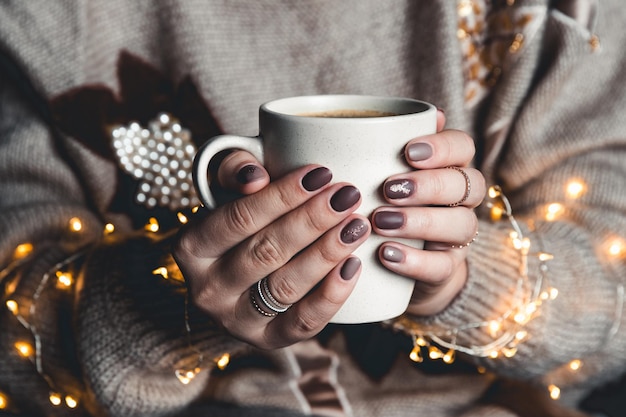 Una mujer con ropa de abrigo de tonos neutros con una guirnalda navideña de rodillas se calienta las manos sobre una taza de esmalte blanco con chocolate caliente y malvaviscos. El ambiente acogedor y navideño.