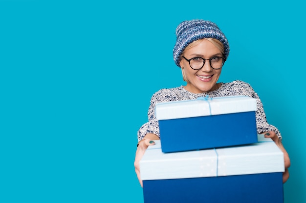 Mujer con ropa azul y sombrero está sonriendo y dando a la cámara cajas presentes mientras usa gafas en una pared azul del estudio