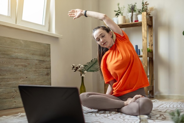 Mujer con ropa brillante en el suelo frente a una computadora portátil está haciendo yoga usando una lección de video