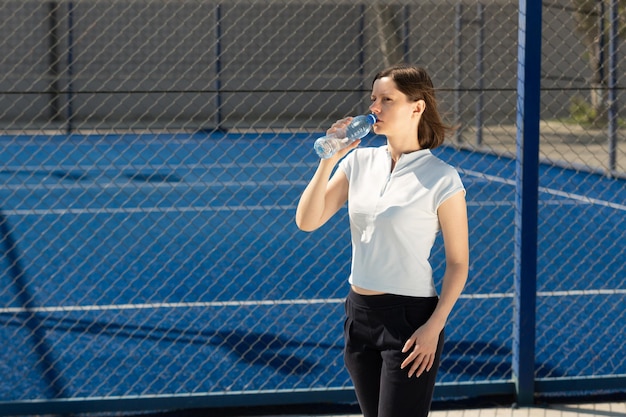 Mujer en ropa deportiva con una botella de agua potable cerca del campo de deportes de tenis