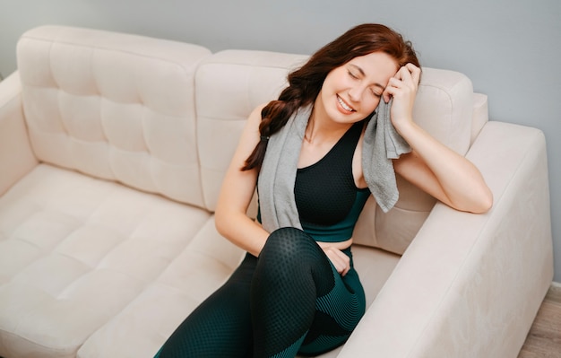 Mujer en ropa deportiva está sentada en el sofá en casa después de yoga con una toalla de sudorconcepto de ...