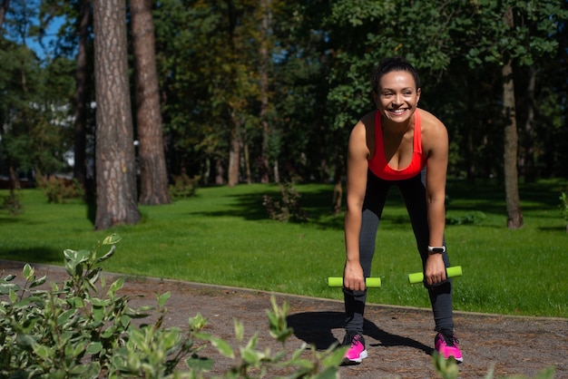 Mujer en ropa deportiva haciendo ejercicios de peso muerto con mancuernas y sonriendo a la cámara