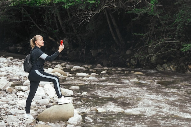 Una mujer en ropa deportiva y con una mochila toma fotos de la naturaleza en las montañas por teléfono