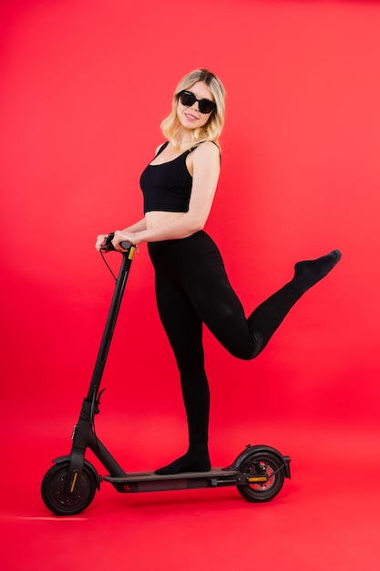 Mujer con ropa deportiva de pie en scooter eléctrico con gafas de sol en fondo rojo y blanco