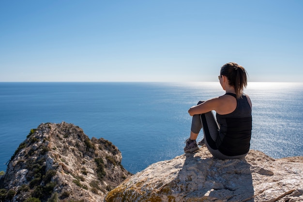 Mujer con ropa deportiva sentada sobre unas rocas observando el paisaje del mar Mediterráneo