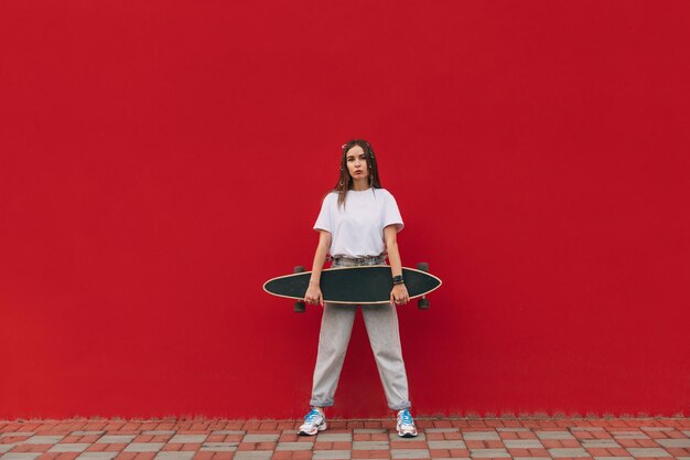 mujer con ropa informal de la calle posando en la cámara con un longboard en las manos sobre un fondo de pared roja