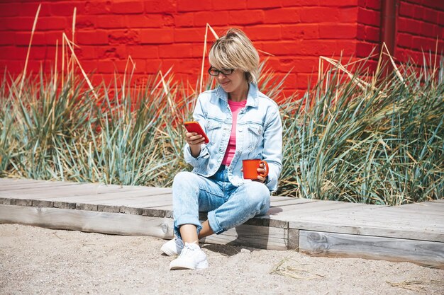 Mujer con ropa informal de mezclilla usando un teléfono inteligente rojo mientras bebe café de una taza roja sentada al aire libre Tecnología de estilo de vida y concepto de red de medios sociales