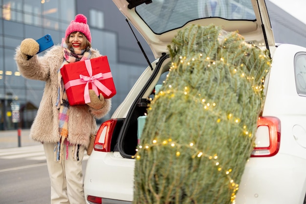 Mujer con ropa de invierno habla por teléfono en línea mientras hace compras para unas vacaciones de invierno en un centro comercial de pie con una caja de regalos cerca de un árbol de Navidad recién comprado al aire libre